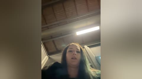 Media: Video of a young woman with long dark hair, wearing a black jacket, captured from a low angle inside a dimly lit room with a wooden ceiling.