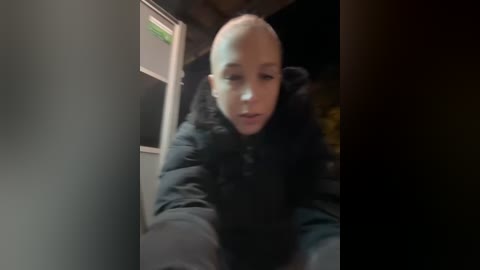 Media: A blurry video of a bald man with light skin, wearing a black puffer jacket, standing in a dimly lit, cluttered room with wooden walls and a refrigerator in the background.