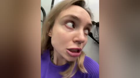 Media: Video of a young woman with fair skin, blonde hair, and a purple sweater, looking surprised and slightly open-mouthed, in a bathroom with a black sink and mirror background.