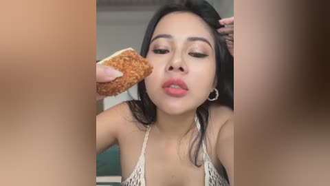 Media: Video of a young Asian woman with long black hair, wearing a white halter top, eating a piece of toast. She has light skin and is holding the toast close to her face. The background is blurred.