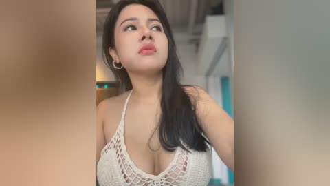 Media: Video of a young Asian woman with long black hair, wearing a beige crochet top, looking upward in a modern, indoor setting with blurred background.