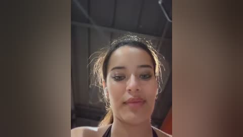 Media: A close-up video of a young woman with fair skin, dark hair pulled back, wearing a black sports bra. She has a neutral expression and is indoors, with a blurred background suggesting a gym or sports facility.