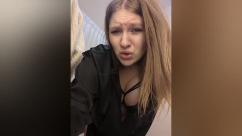 Media: A video of a young, fair-skinned woman with long, straight blonde hair, wearing a black blouse with a visible bra strap, making a pouty face while partially obscured by a dark object.