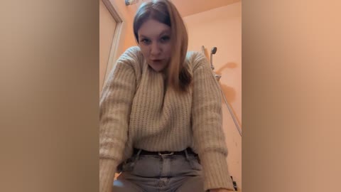 Media: Video of a young Caucasian woman with light skin and long, straight brown hair, wearing a cream knitted sweater and high-waisted jeans, leaning forward in a hallway with beige walls.