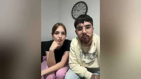 Media: Video of a young woman with light skin, brown hair, and a black top, and a man with dark skin, short hair, and a Nike sweatshirt, sitting on a bed, with a clock on the wall.