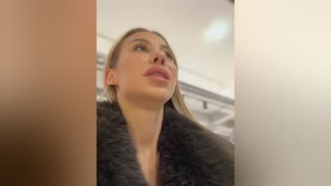 Media: Video of a light-skinned woman with straight blonde hair, wearing a black fur coat, gazing upwards with a serious expression. Background features a blurred indoor setting with neutral tones.