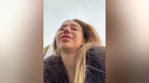 Media: Video of a smiling woman with long blonde hair, wearing a dark fur coat, taken from a low angle, with a blurred, cloudy sky background.