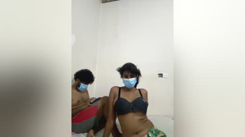 Media: Video of a woman with medium brown skin, wearing a blue surgical mask and black bra, sitting on a red and green blanket, reflected in a mirror.