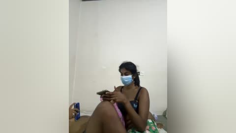 Media: Video of a young South Asian woman with dark skin and medium-length black hair, wearing a blue surgical mask, sitting on a bed in a sparsely furnished room with white walls. She has a phone in her hand.