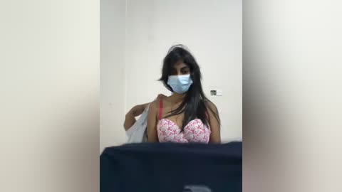 Media: A video of a South Asian woman with long black hair, wearing a floral bra, blue face mask, and a white shirt, standing in a plain white room.