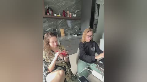 Media: A video shows two young women with fair skin and light hair, wearing casual clothes, seated at a modern kitchen counter with a marble backsplash and wine bottles.