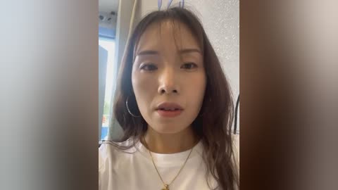 Media: Video of a young Asian woman with long, dark hair and light skin, wearing a white shirt and gold necklace, standing indoors against a blurred background.