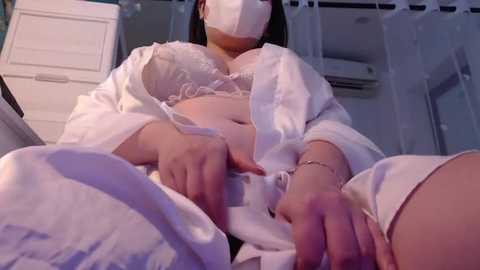 Media: Video of an Asian woman in white lingerie and robe, seated with hands on her stomach, wearing a white mask, in a dimly-lit, modern room with white walls and a white cabinet.