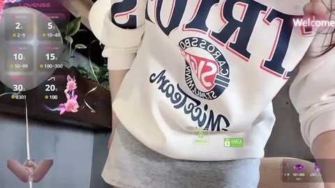 Media: Video of a woman wearing a white \"Tigers\" sweatshirt, standing indoors with a pink flower in the background and a virtual reality display showing a hand.