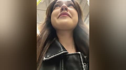 Media: Video of a young woman with fair skin and long brown hair, wearing a black leather jacket, looking upward with a slight smile, in an indoor setting with a white ceiling.