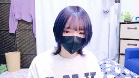 Media: Video of an East Asian woman with straight black hair and bangs wearing a black face mask, white shirt, and a patterned blanket. Background includes a white curtain, wicker screen, purple jacket, and a blue chair.