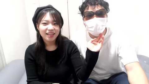 Media: Video of an East Asian woman with long black hair, wearing a black hat and black long-sleeve shirt, smiling. Beside her, an Asian man with curly black hair, wearing a white mask, sunglasses, and a white t-shirt.