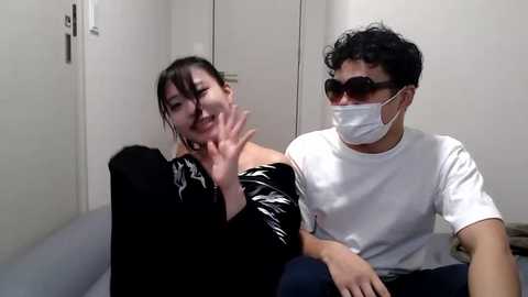 Media: Video of two people in a plain room: a woman with dark hair and black clothing, and a man with curly hair, wearing a white T-shirt, mask, and sunglasses.