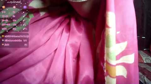 Media: Video of a close-up of a person wearing a pink saree with a floral design, partially obscured by a pink handkerchief. The background features a dimly lit room with a dark wall and a bed with a blanket.