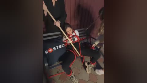 Media: Video of a woman in black bondage gear, bound with red rope, seated on a black bench, surrounded by a dimly lit, rustic room with brick walls.