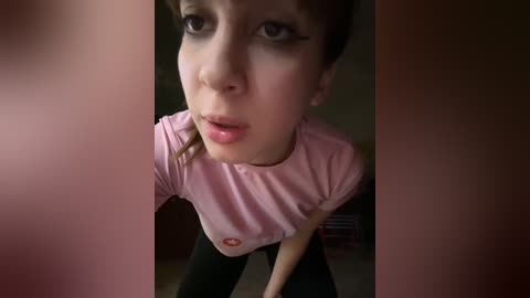 Media: A video of a young, fair-skinned woman with brown hair, wearing a pink t-shirt and black leggings, leaning forward, slightly out of focus.