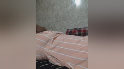 Media: Video of a sleeping person, covered in a light pink, striped blanket, with a plaid pillow in the background. The room has a textured, beige wall and a dim light source overhead.