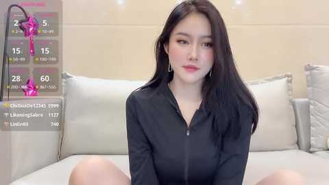 Media: Video of a young Asian woman with long black hair, sitting on a white bed, wearing a black zip-up hoodie, in a modern, minimalist bedroom.