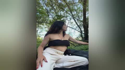 Media: Video of a young woman with long dark hair, wearing a black strapless top and white high-waisted pants, sitting outdoors among trees.