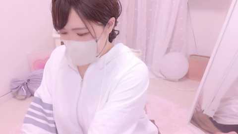 Media: A video of an Asian woman with dark hair in a bun, wearing a white face mask, white lab coat, and a gray-striped sleeve, sitting on a bed with white sheets and a pink blanket in a softly lit room.