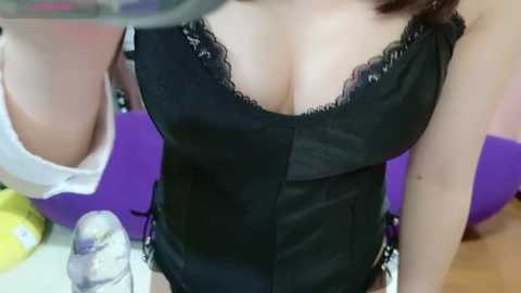 Media: Video of a woman in a black corset with lace trim, medium breasts, fair skin, and brown hair partially visible. Background includes purple and white objects.