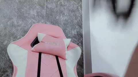 Media: Video of a pink gaming chair with white accents and a small black pillow, positioned on a textured, grayish-brown tiled floor. The background shows a blurred white wall with a faint outline of a person.