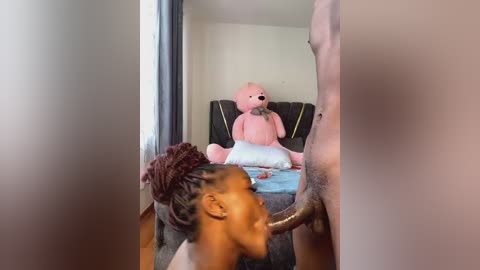 Media: Video of a dark-skinned woman with braided hair performing oral sex on a light-skinned man. A stuffed pink duck is on a bed in the background.