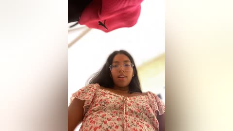 Media: A video of a young woman with medium-brown skin and long black hair, wearing glasses and a floral blouse, looking up at a pink object above her head.