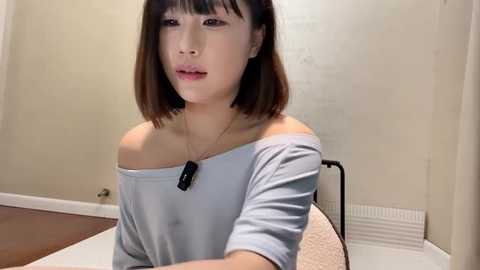 Media: Video of a young Asian woman with straight, shoulder-length brown hair, wearing a light blue off-shoulder top, sitting on a chair in a beige-walled room with a radiator.