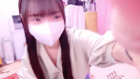 Media: A video of an Asian woman with straight, long brown hair wearing a white face mask, beige blouse, and making a selfie gesture in a brightly lit room with pink and white decor.