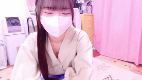 Media: A video of an Asian woman with long brown hair and fair skin, wearing a white face mask, a beige robe, and kneeling on a white rug in a pink room with a wooden dresser and white curtains.