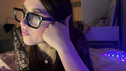 Media: Video of a young woman with fair skin, long brown hair, and black-rimmed glasses, lying on a bed, wearing a black lace top, looking thoughtful. Background features a dimly lit bedroom with a white dresser and a blue string of lights.