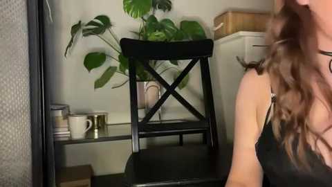 Media: A video of a woman with long, wavy brown hair, wearing a black tank top, sitting on a black wooden stool with a cross design. The background features a potted monstera plant, a beige box, and a shelf with white and gold mugs.