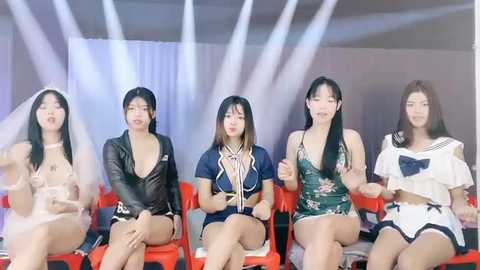 Media: Video of five Asian women, diverse in age and appearance, sitting on red chairs under bright stage lights. They wear revealing, colorful, and patterned outfits, some with skimpy tops and shorts. Background shows a simple stage setup.