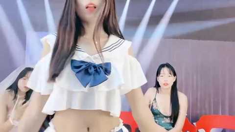 Media: Video of three Asian women: one in a white crop top with a blue bow, another in a green top, and a third in a blue top, all seated on red chairs in a modern room with white walls and a mirror.