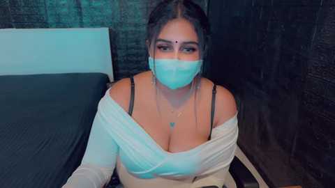 Media: Video of a South Asian woman with medium skin tone, wearing a white off-shoulder top, black bra, and blue mask, sitting on a bed with a dark headboard.