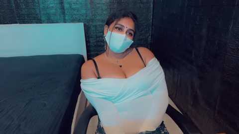 Media: Video of a curvy South Asian woman with medium skin tone and dark hair, wearing a white off-shoulder top, black bra, and a face mask, seated on a bed in a dimly lit room.