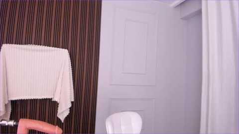 Media: A video shows a white hand holding a beige towel draped over a brown, vertical-striped wall. A white chair is partially visible in the background. The room is minimalist and modern with a white door and curtain.