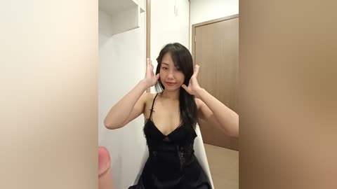 Media: Video of an Asian woman with long black hair, wearing a black strapless dress, playfully pulling her hair. She sits in a beige-walled room with wooden doors and a mirror.