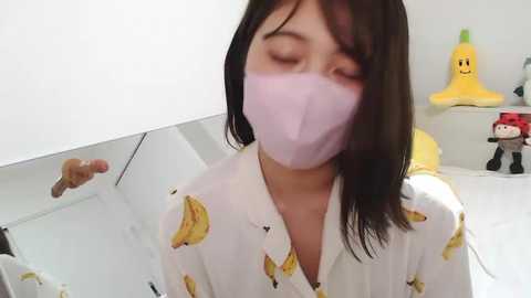 Media: Video of a young Asian woman with long black hair, wearing a white shirt with banana pattern, face mask, and closed eyes. Background includes a white wall, a yellow banana plush toy, and a red plush toy.