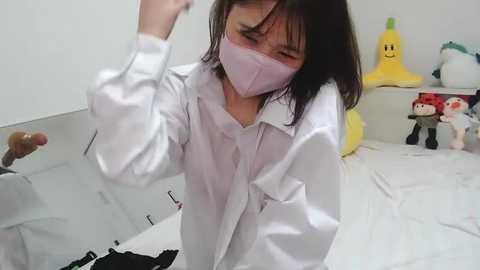 Media: Video of a woman with dark hair, wearing a white lab coat, mask, and glasses, dancing joyfully in a brightly lit room with yellow toys and plushies.