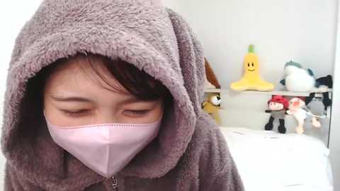 Media: Video of a young Asian child with light brown skin, wearing a gray plush hooded robe with a pink face mask, eyes closed, standing in a bedroom with colorful plush toys on shelves in the background.