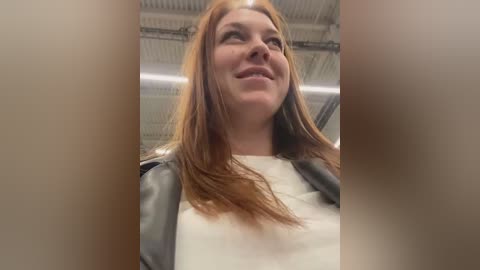 Media: Video of a smiling, fair-skinned woman with long, straight, auburn hair, wearing a white top and a gray leather jacket. The background features an industrial setting with exposed metal beams and fluorescent lighting.