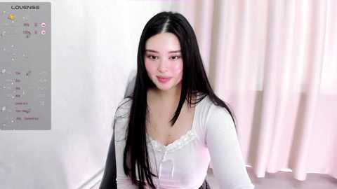 Media: Video of a young East Asian woman with long black hair, fair skin, and wearing a white blouse, sitting indoors with pale curtains in the background.