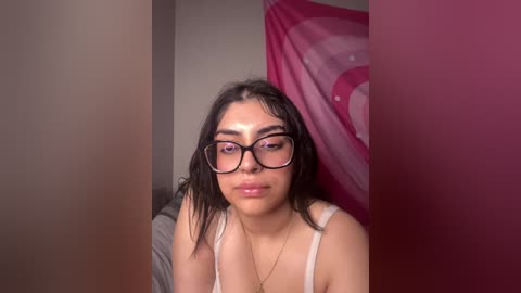 Media: Video of a young woman with medium skin tone, dark hair, and glasses, wearing a white tank top, in a dimly lit room with pink decor.